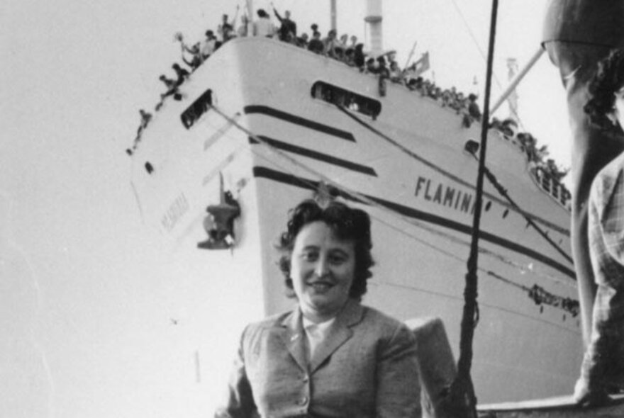 Italian woman standing in front of a ship with passengers crowding the prow deck.
