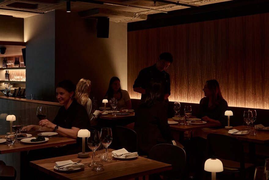 A dark and moody restaurant setting with with patrons seated at wooden tables, all enjoying glasses of wine.