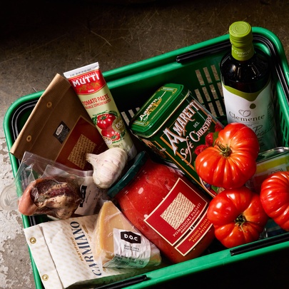 A green plastic grocery basket contains tomato paste, cookies, oil, and fresh tomatoes.