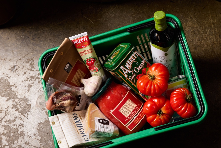 A green plastic grocery basket contains tomato paste, cookies, oil, and fresh tomatoes.