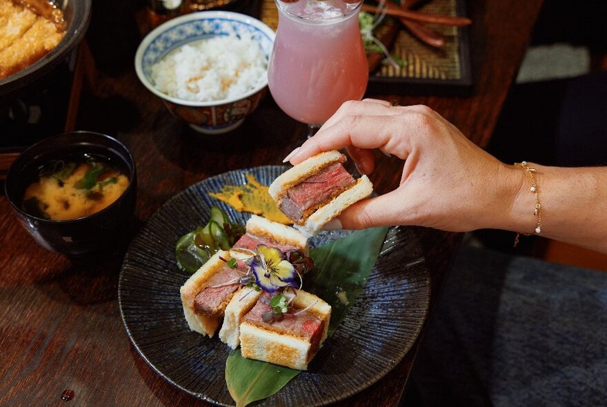 A hand picking up a piece of katsu sando from a delicately plated serving, with a side of white rice, miso soup and a pink grape cocktail.