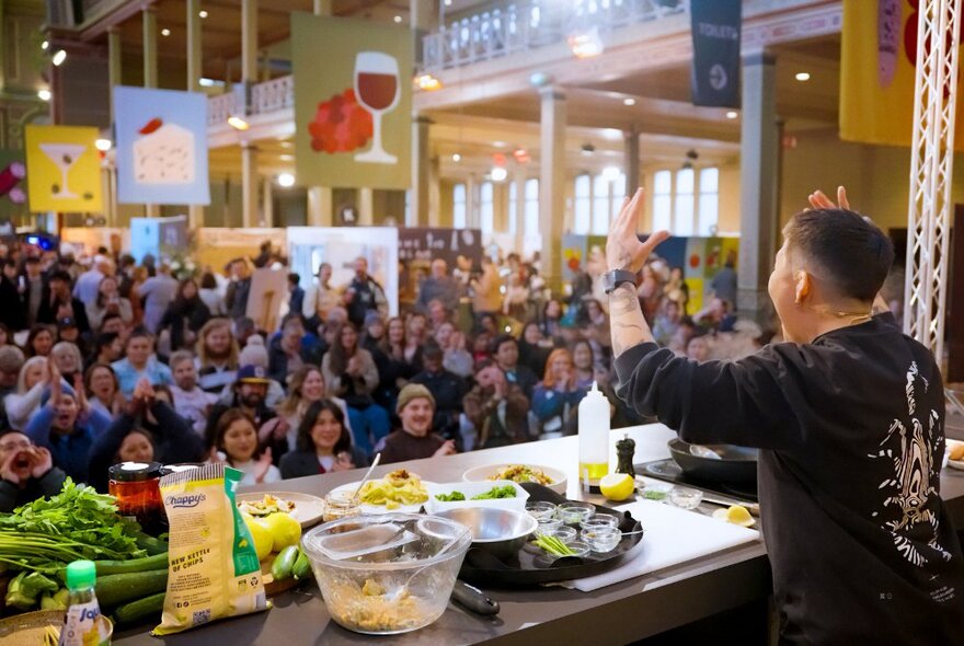 A chef hosting a cooking demo at a table, with a crowd of people watching, inside the Royal Exhibition Building.