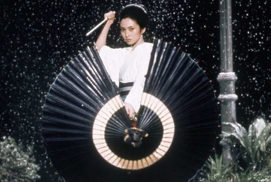 A still from the movie Lady Snowblood depicting a woman in a white dress holding a sword in her hand, standing behind a large black parasol outdoors.