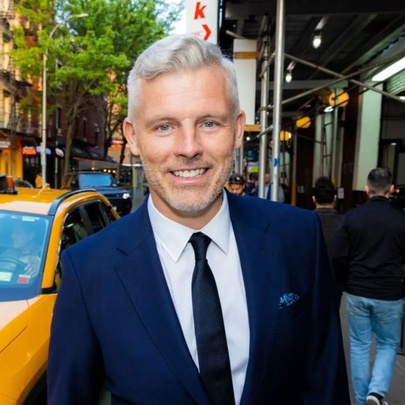 A smiling middle aged man with white hair wearing a navy coloured suit and tie, standing outdoors in a busy city street with a yellow taxi behind him.