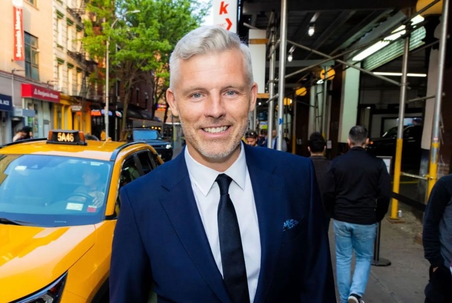 A smiling middle aged man with white hair wearing a navy coloured suit and tie, standing outdoors in a busy city street with a yellow taxi behind him.