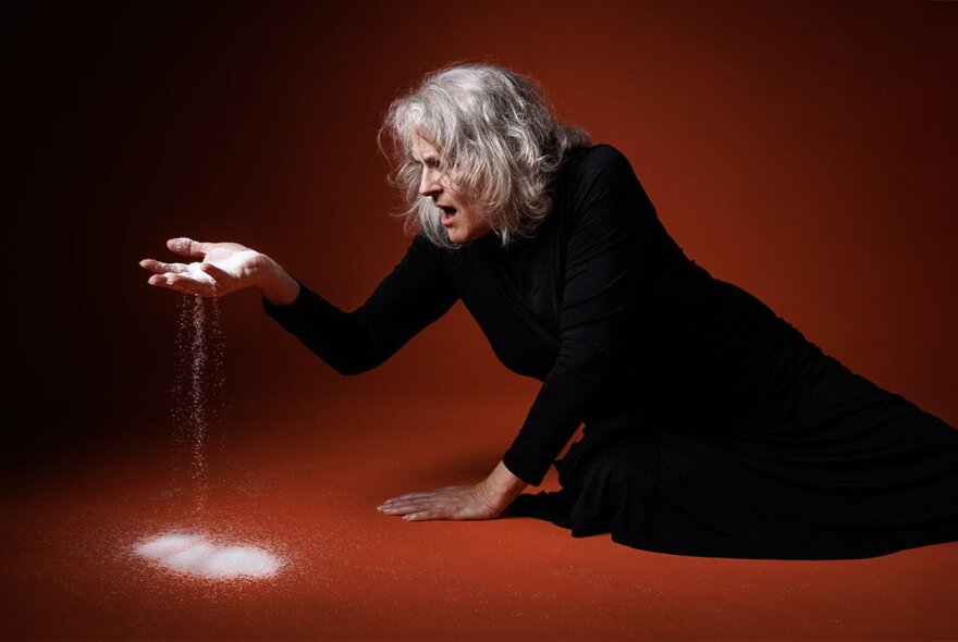 A woman in black on the floor  of a red room sifting sand through her fingers looking anguished. 