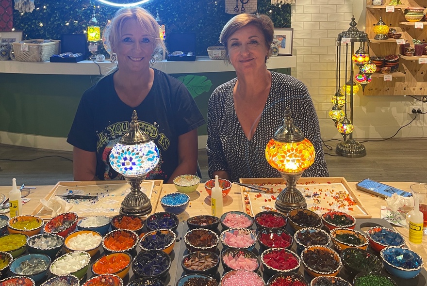 Two women seated at a craft table at a workshop session in a studio, with multiple bowls of coloured glass and two small mosaic lamps in front of them.