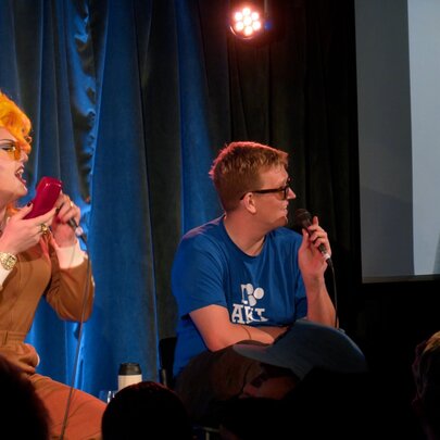 Two comedians seated on a stage in front of an audience, interacting with someone on a large projected screen on stage next to them.