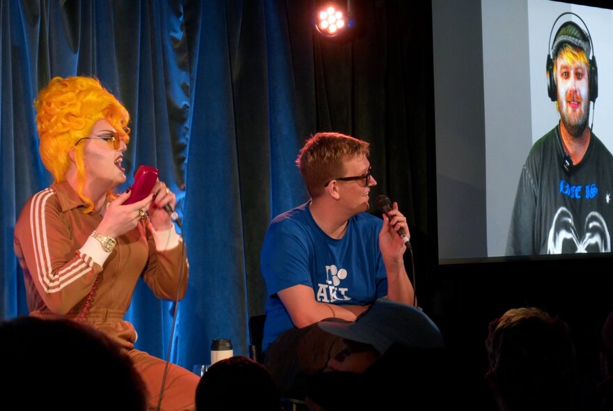 Two comedians seated on a stage in front of an audience, interacting with someone on a large projected screen on stage next to them.