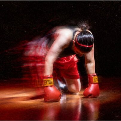 A scene from the play Bitch Boxer, featuring a performer on all fours, in red boxing shorts, singlet and boxing gloves captured mid-motion in a long exposure motion blur.