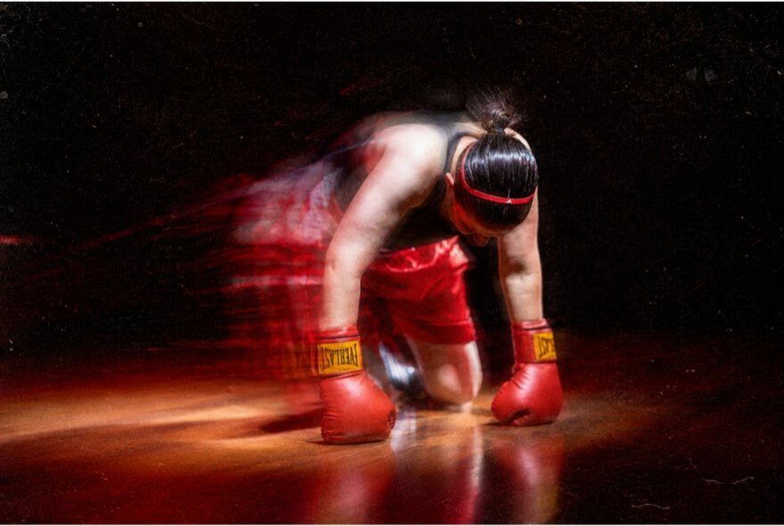 A scene from the play Bitch Boxer, featuring a performer on all fours, in red boxing shorts, singlet and boxing gloves captured mid-motion in a long exposure motion blur.
