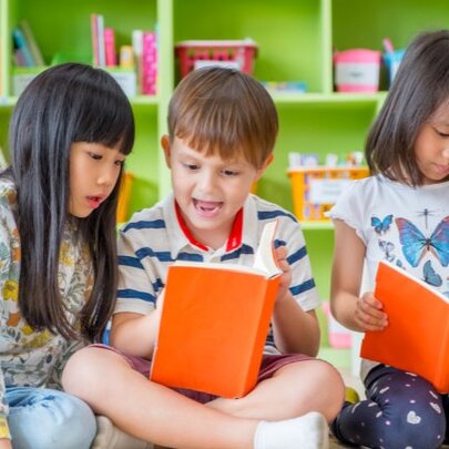 Three young children looking at open books they hold, participating in activities in a library setting.