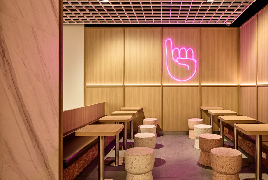 A seating area in a cafe with a pink neon wall sign of a hand holding up a pinky finger.