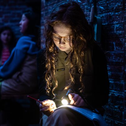 A young girl with long hair holding a light in her hands while resting a book on her lap. 