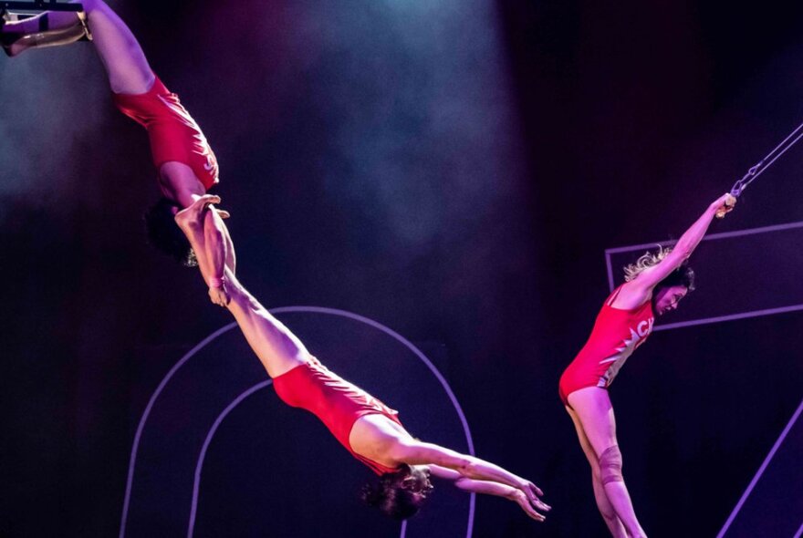 Trapeze artists swinging through the air in red leotards.