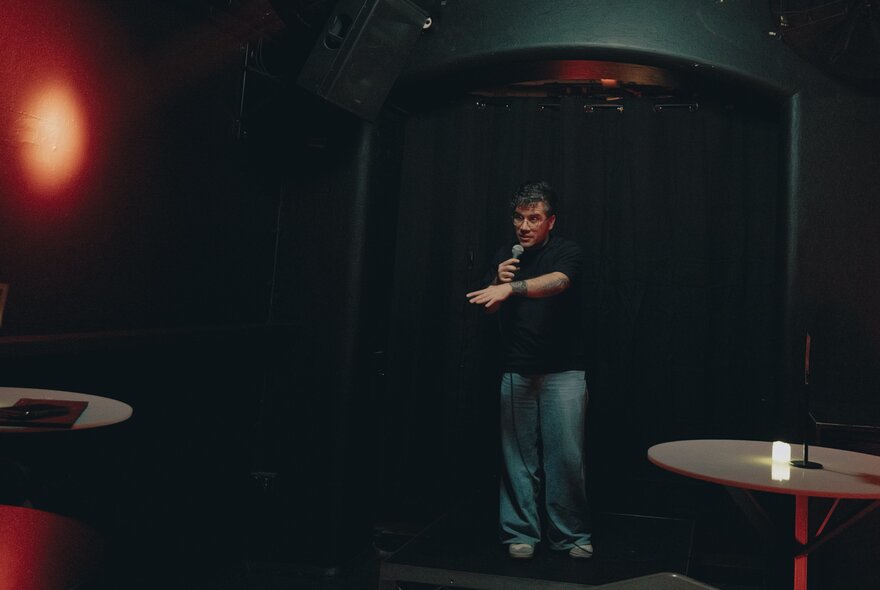 A dark image of a comedian on stage in a black T-shirt and jeans.