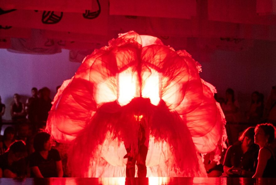 A large red illuminated object, possibly a human figure, comprised of layers of fabric and pillars of light, in a nightclub setting with people just visible at the edges of the picture.