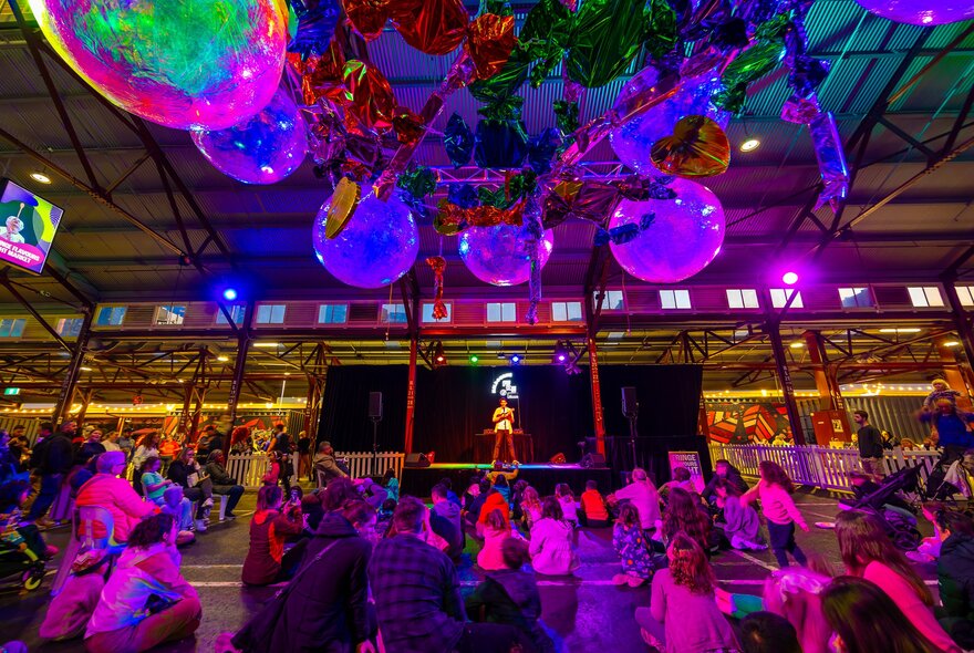 A person is playing the ukulele on a stage to a crowd of children. There are large circular colourful decorations hanging above them. 