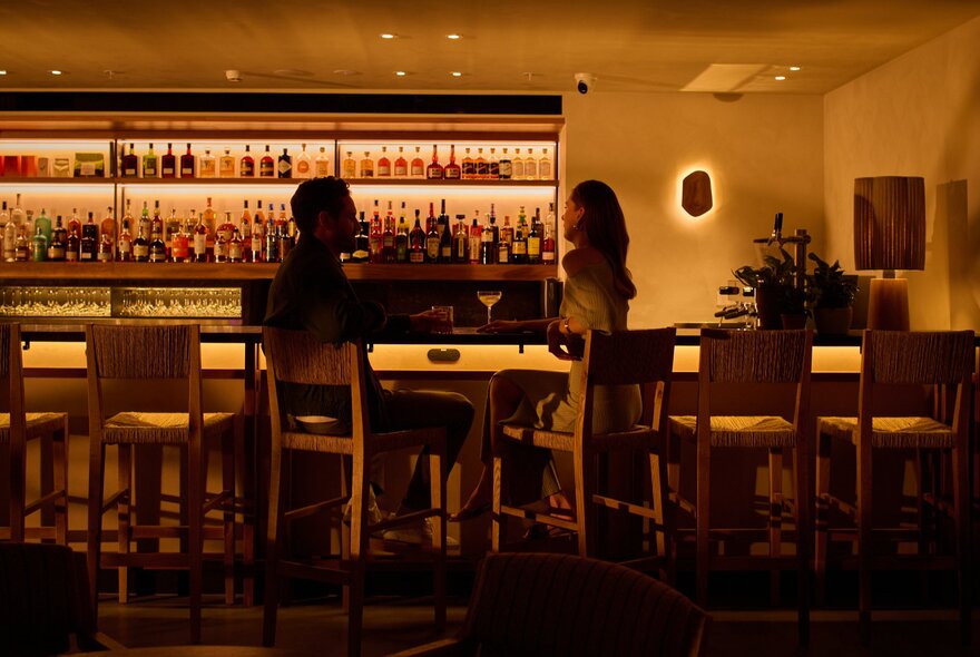 The bar Upstairs at 1 Hotel Melbourne, shelves of bottles on the rear wall behind the bar, soft wall lights, and high bar stools.