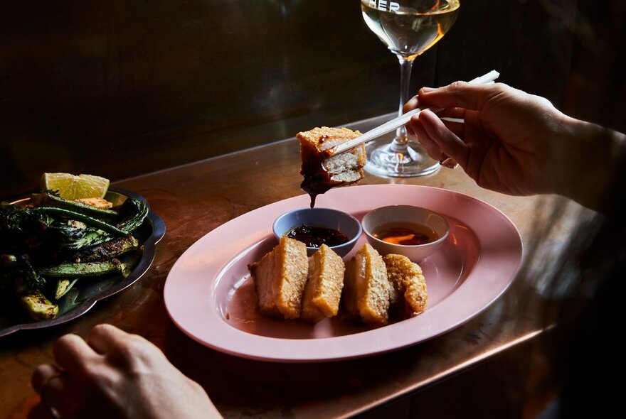 A person holding a piece of Thai pork belly in chopsticks, over a pink oval dish with more slices of pork belly.