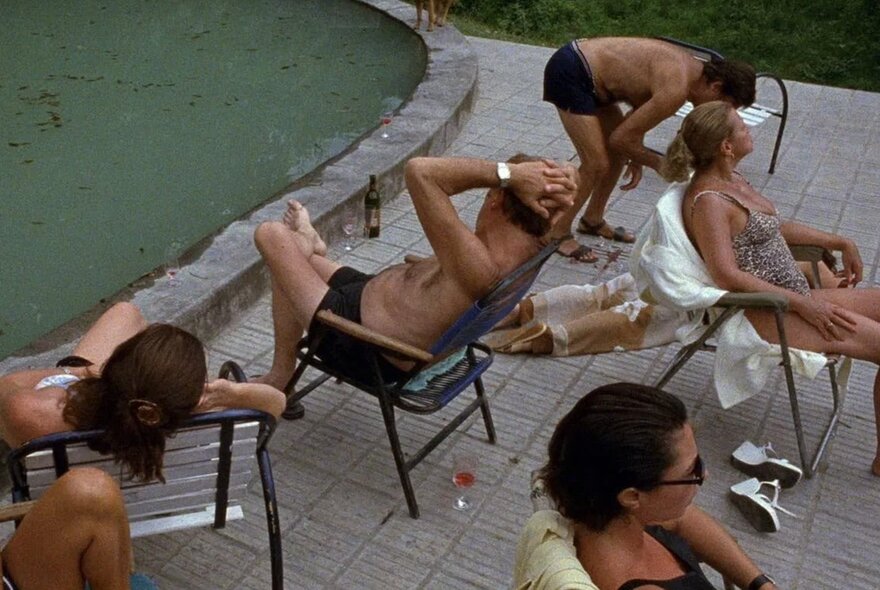 A still from a film, with people in bathing costumes seated on chairs around a swimming pool.