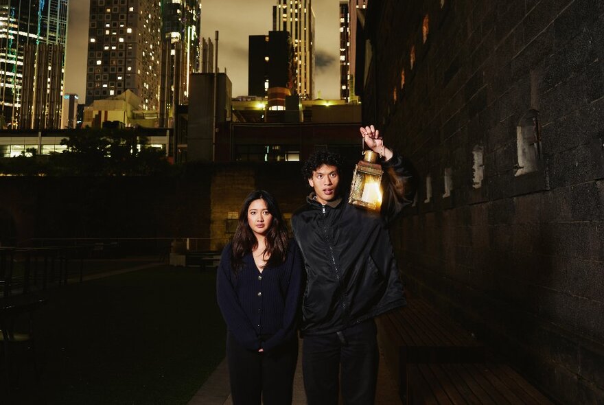 Two people walking in a darkened part of the city, one holding up a lantern.