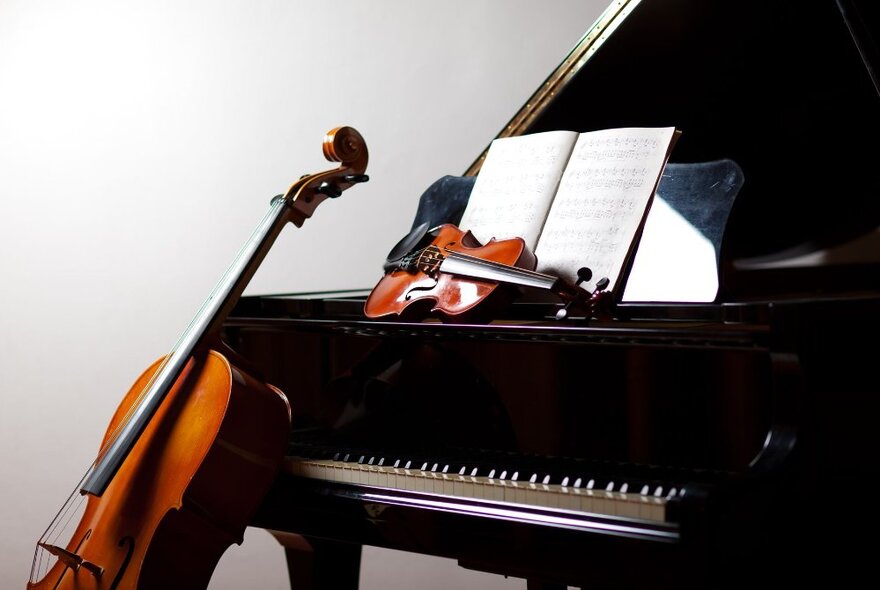 A cello leaning against a black grand piano, and a violin resting on its side in front of the sheet music rest on the piano.