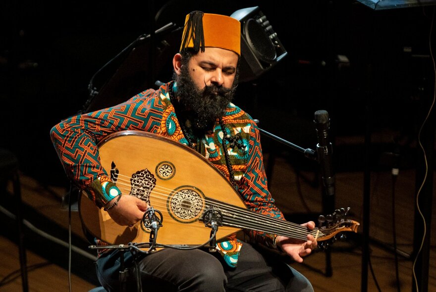 Joseph Tawadros seated on a chair, wearing a patterned shirt and a fez on his head, playing the oud.