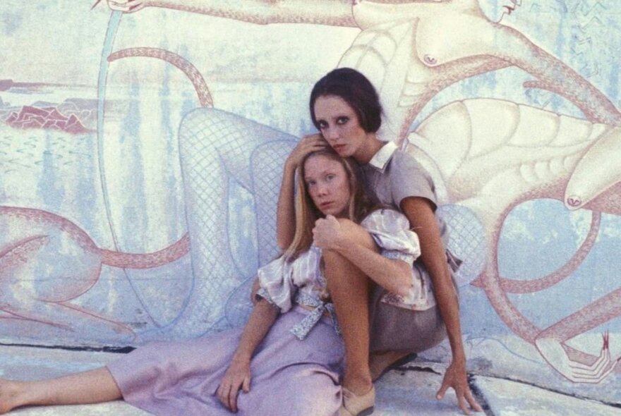 Two young women entwined in an embrace, both wearing tones of lavender in front of a similar-toned wall mural.