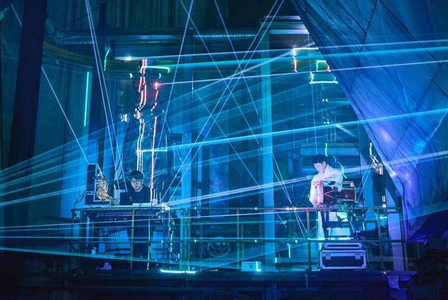 An immersive sound and light show on stage, with blue lasers and three people operating technical electronic equipment.