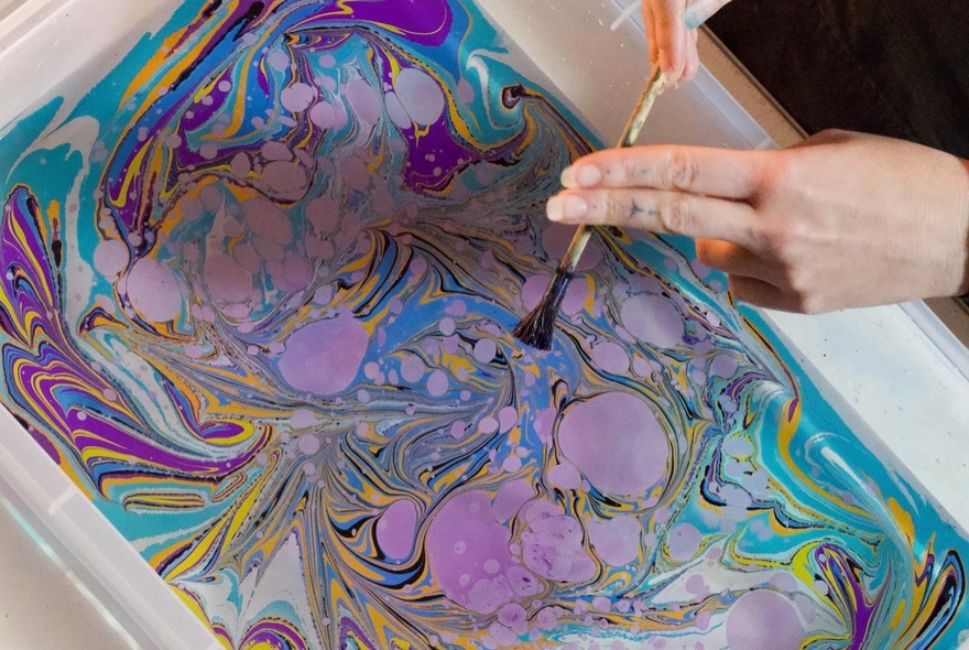 Hand holding a brush over marbled pattern in a water bath.