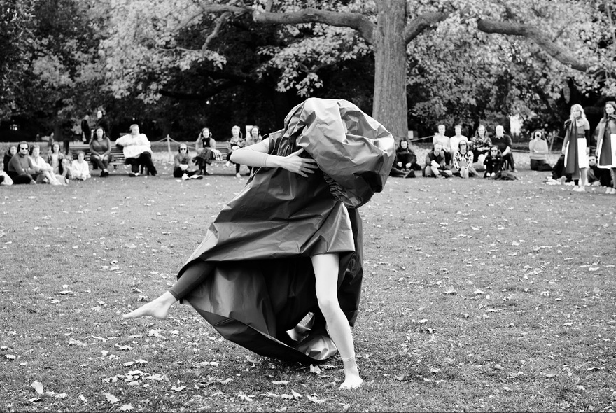 Dancer performing on grass in front of trees and a circle of onlookers, her body and head wrapped in a bag.