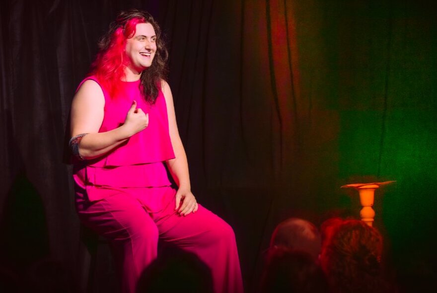 Comedian with dyed hair seated on a chair on stage during a performance, wearing cerise outfit.