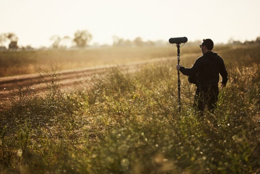 A film sound technician holding equipment in a field with filtered sunlight. 
