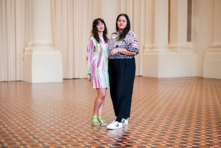 Two women stand together in an empty room with white columns and a tessellated tile floor. 