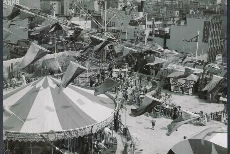 Black and white photo of a carnival held on a rooftop featuring a Ferris wheel, carousel and bunting flags 