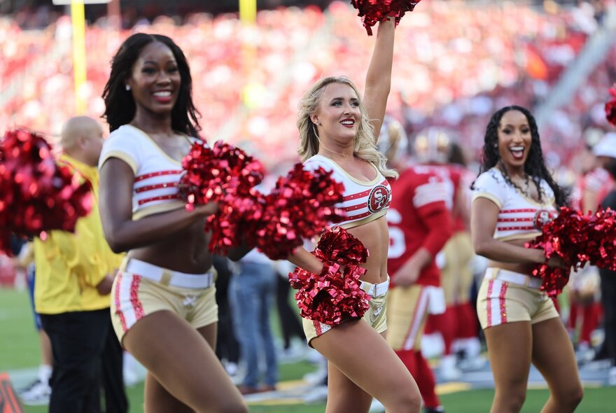 Three 49ers cheerleaders holding shiny red rosettes in front of arena stands.
