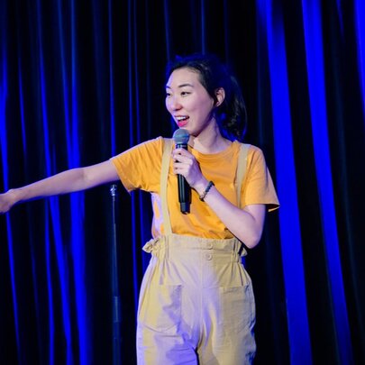 A woman in overalls performing stand up comedy holding a microphone with one hand outstretched. 