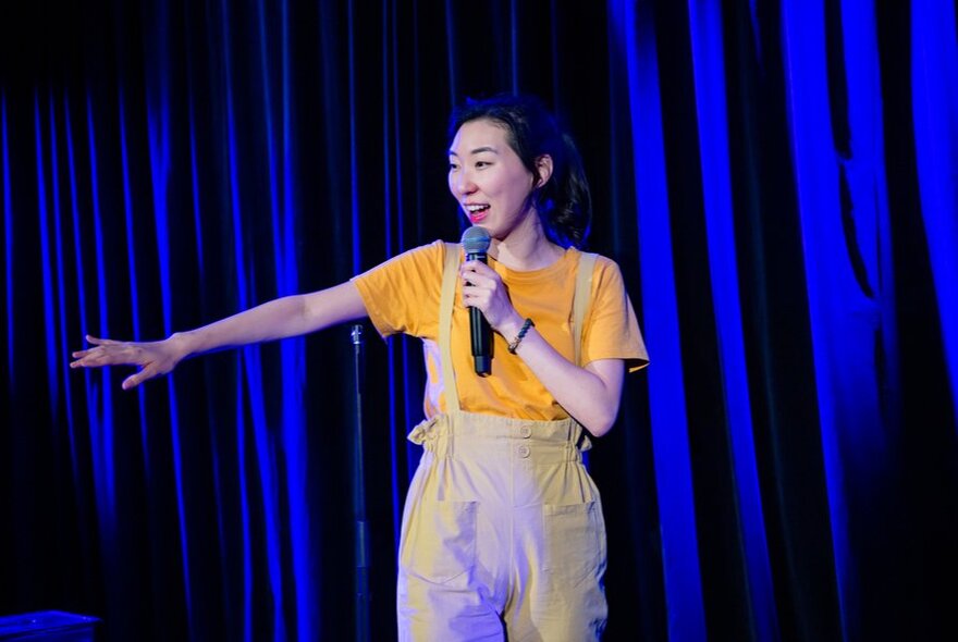 A woman in overalls performing stand up comedy holding a microphone with one hand outstretched. 