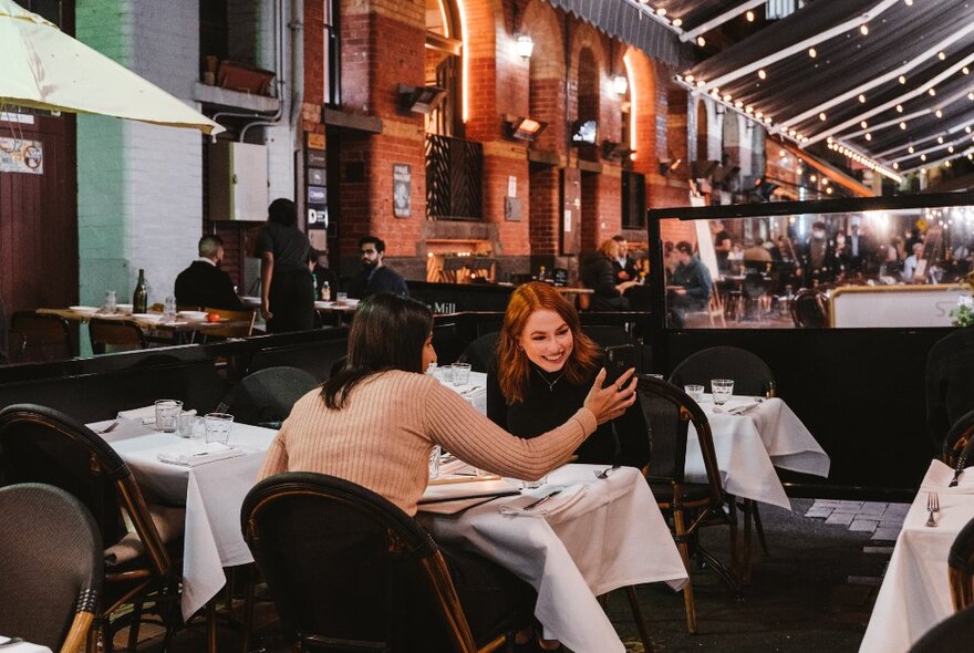 Diners seated at an outdoor table with white tablecloth in a laneway with overhead outdoor lighting.