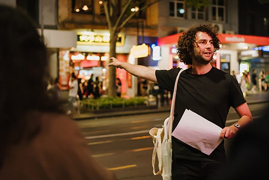 A man holding a piece of paper with a satchel on his shoulder, pointing his arm towards brightly lit shops on the other side of the road behind him.