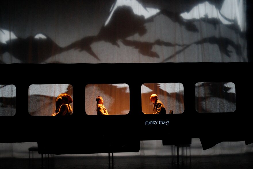 Looking at a stage set of a black building with people visible in the windows and mountains in the background. 