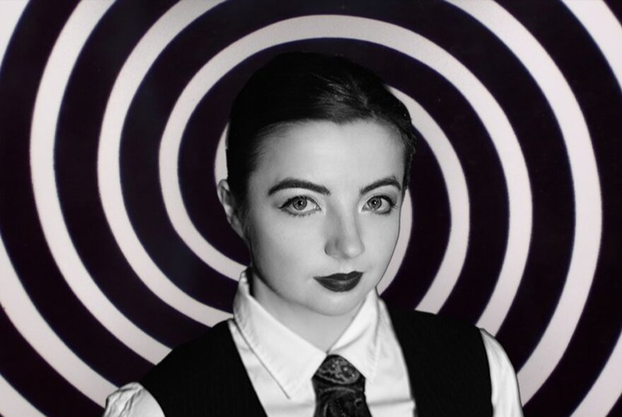 A black and white image of a woman wearing a waistcoat and tie, in front of a black and white spiral.