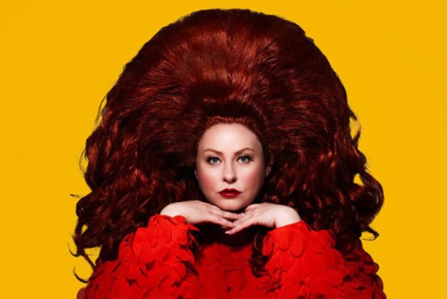 Comedian Amy Glenhill with comically enormous red hair, a red top and a yellow background. 