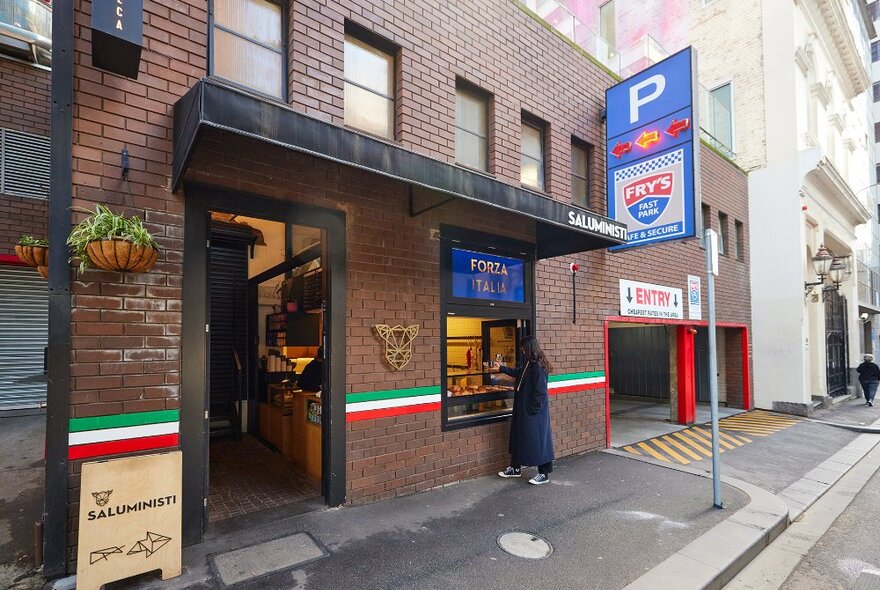 A woman ordering from an Italian themed cafe window, next to a carpark with a large blue P sign.