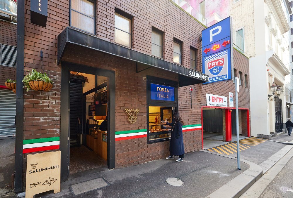 A woman ordering from an Italian themed cafe window, next to a carpark with a large blue P sign.