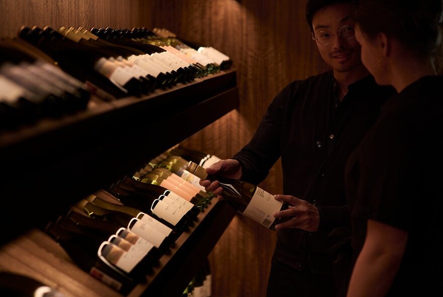 A wine sommelier presenting a bottle of wine to a patron, shelves of wine bottles on the left.