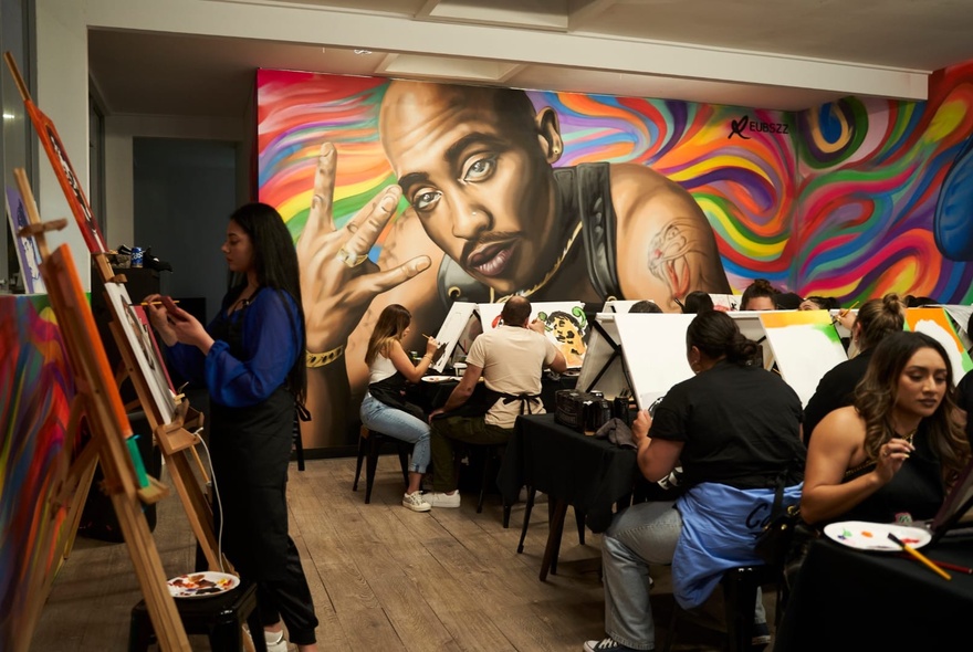 Several rows of people painting on canvases in a room with large, wall murals.