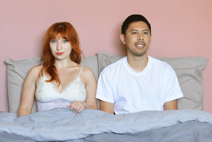 A man and a woman sitting up in bed, he is smiling and she's looking slightly peeved. 