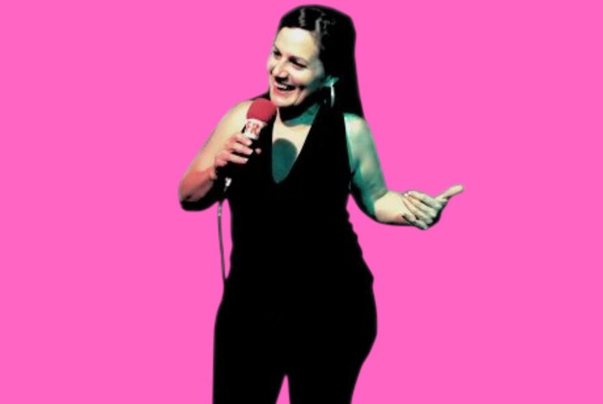 Comedian smiling while holding a microphone, one arm raised, wearing a black pantsuit against a pink background.